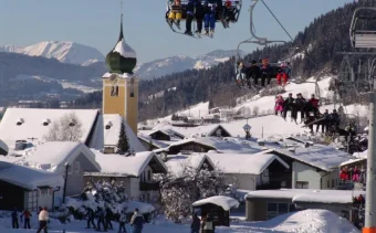 Skiers are carried in chair lifts with an onion shaped domed church and the pretty alpine town of Westendorf, Austria in the background