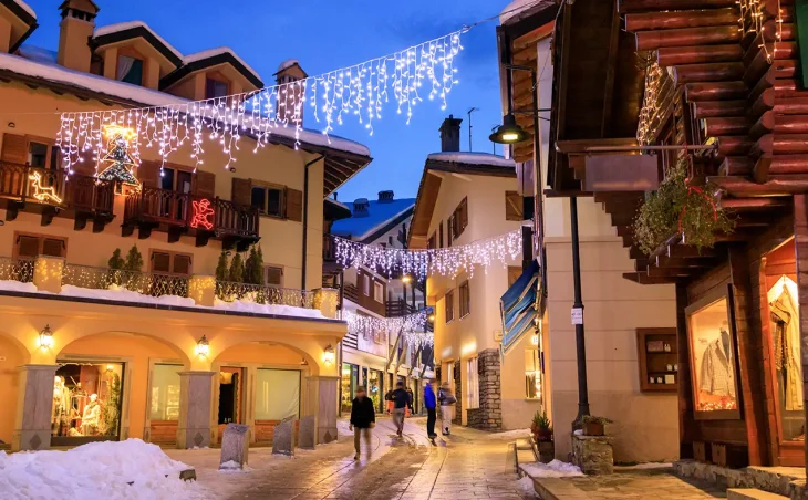 A nighttime scene of the pretty village of Courmayer, Italy. Christmas lights decorate the streets with shoppers beneath.