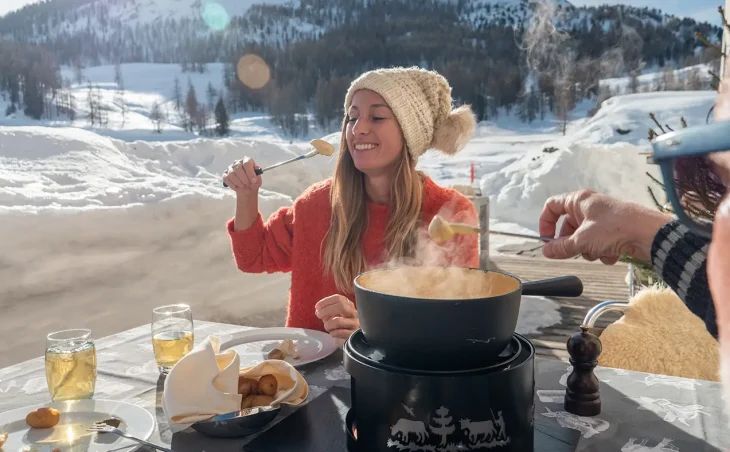 A lady wearing a wooly hat enjoys a Swiss fondue meal on the ski slopes