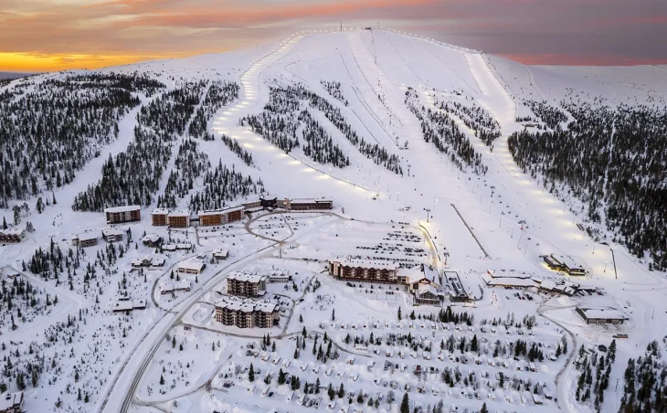 An aerial photograph of the Ylläs ski slopes in Finland. The sun is low turning the sky a shade of orange. Floodlights gently illuminate the pistes. Trees line the edges of the slopes. At the foot of they mountain is Ylläs resort, a great location for a ski holiday in Finland!