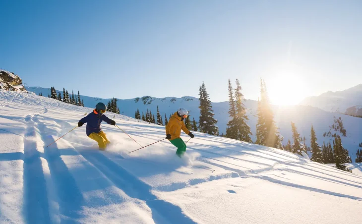 Two skiers in bright outfits enjoying a ski holiday in Austria. They are carving turns in powder snow as the sun shines brightly for across the Austrian Alpine mountains.
