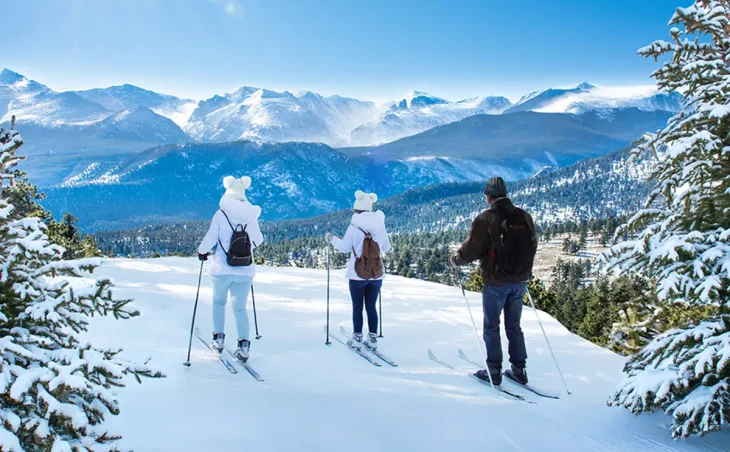 Two female skiers with white pompom hats and one male skier look out over a magnificent Mountain View on a ski holiday in the United States of America